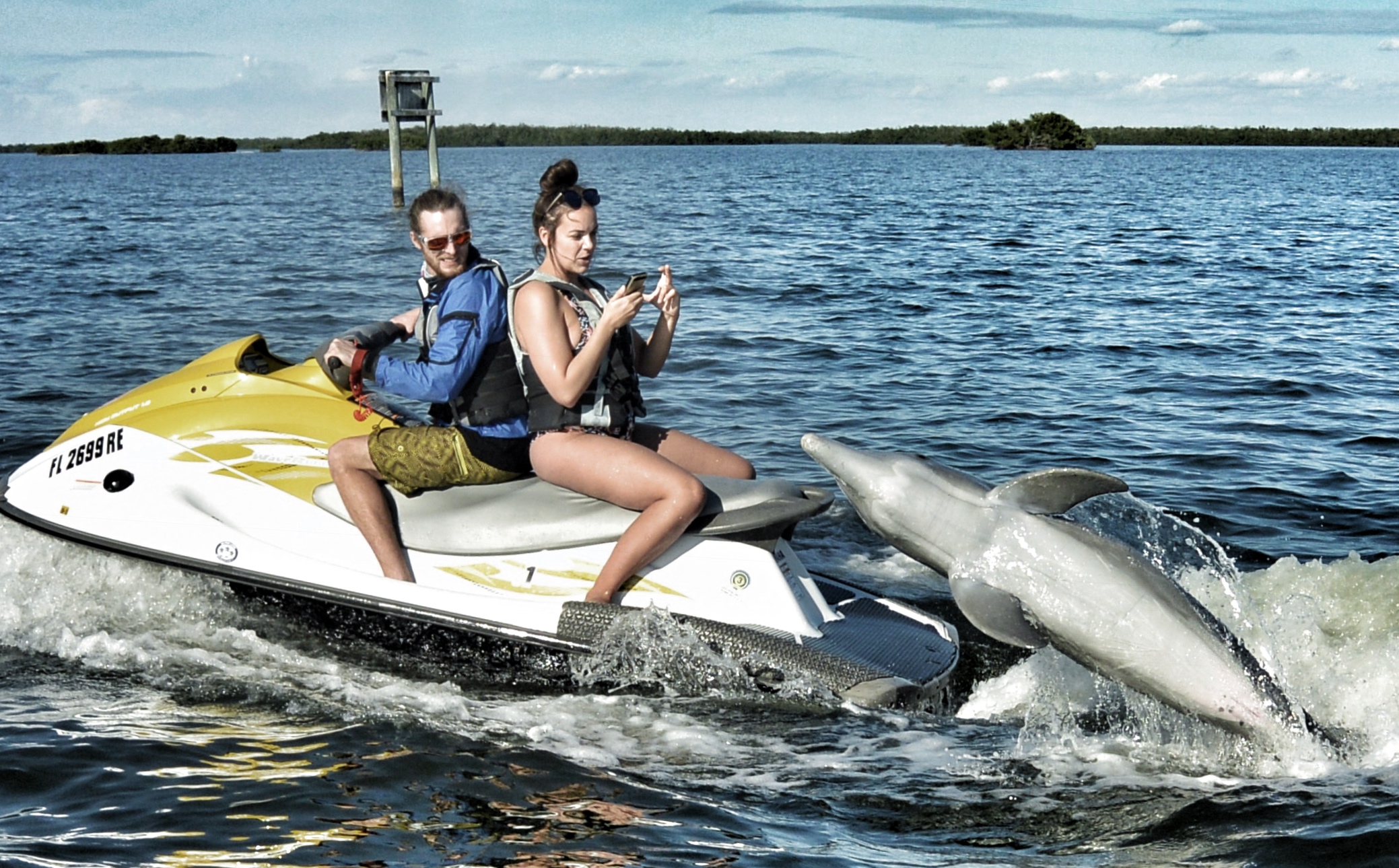 Girl taking picture of dolphin jumping behind the jet ski she is riding on